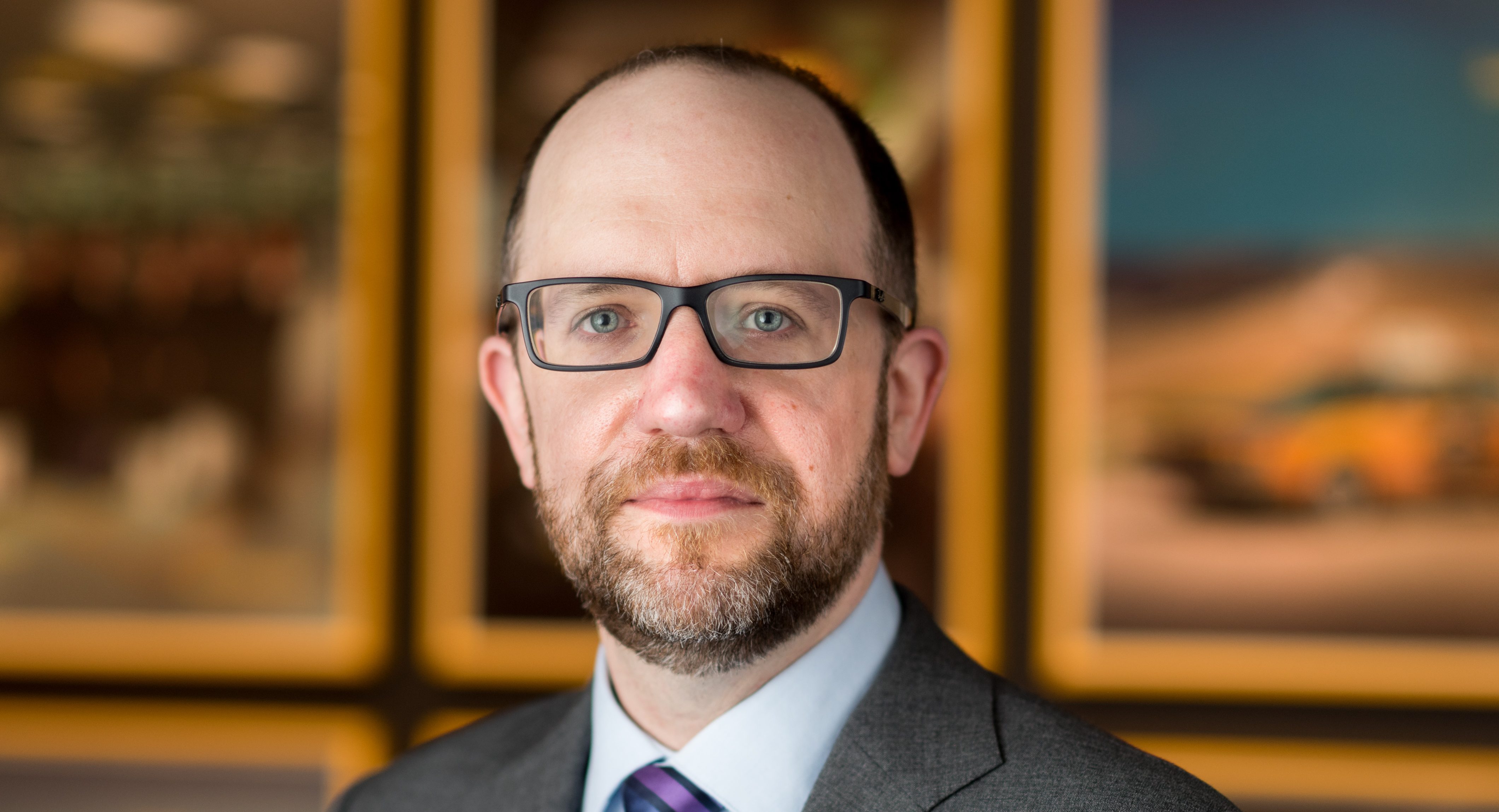 Headshot of a light-skinned man with glasses and a suit in front of a wall with several artworks.