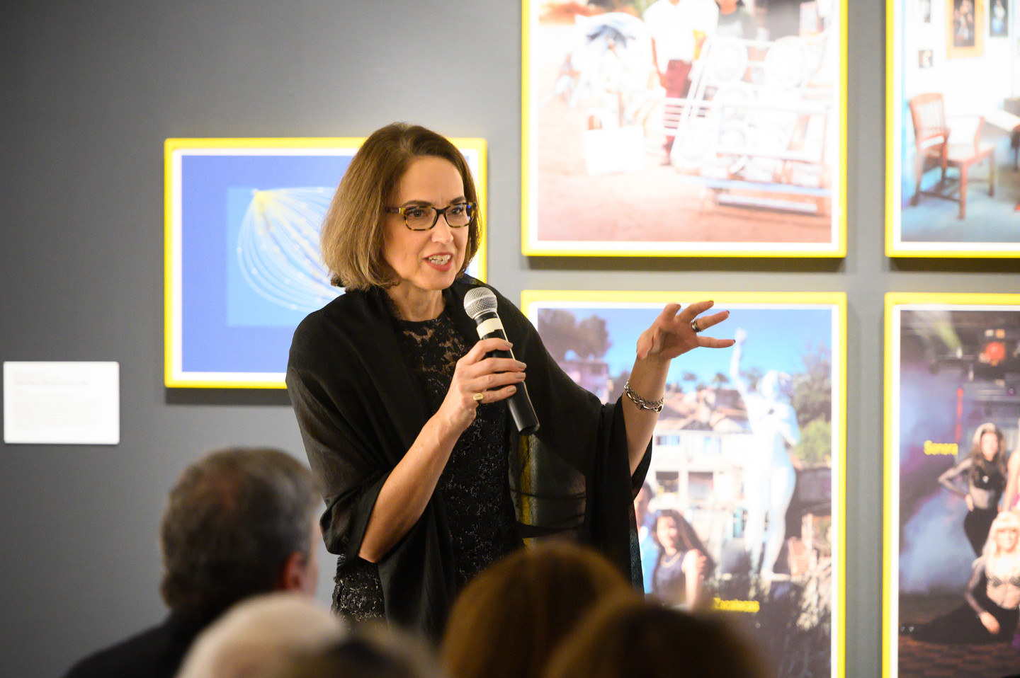 A woman with light skin and dark hair is speaking into a microphone in front of a wall with multiple photographs in bright yellow frames.