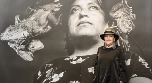 A woman in a fedora and a long black dress poses in front of a large black-and-white photograph of a woman wearing a floral dress and a large headdress who is standing, looking upward.