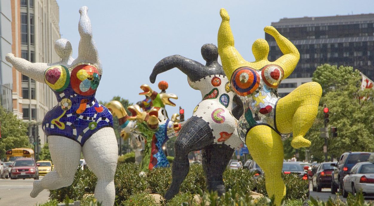Three abstract outdoor sculptures of voluptuous figures covered in bright patterns and dancing with outstretched arms.