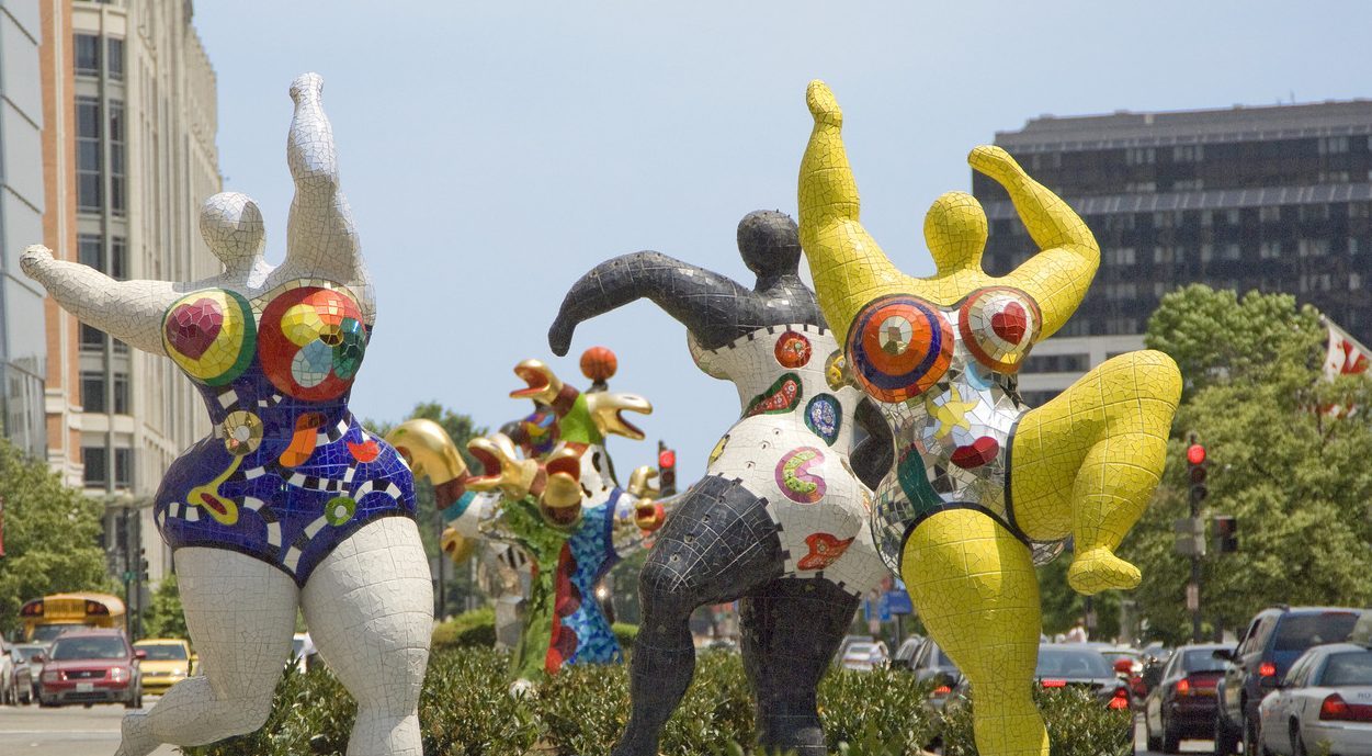 Three abstract outdoor sculptures of voluptuous figures covered in bright patterns and dancing with outstretched arms.