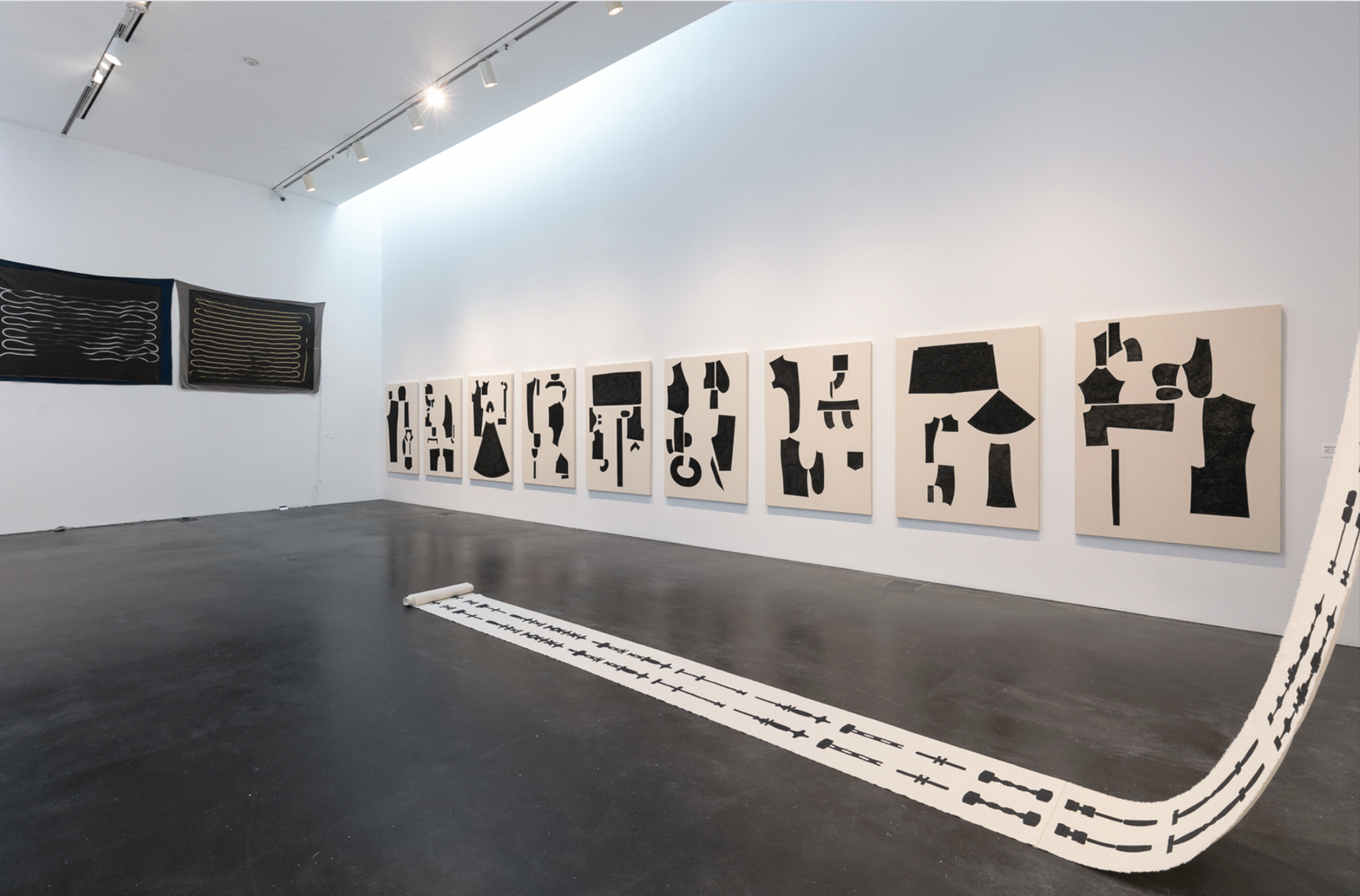 An installation in an industrial gallery space featuring abstract black-and-white prints on one wall and a long white scroll atop which symbols in black are etched. It extends into the middle of the room.