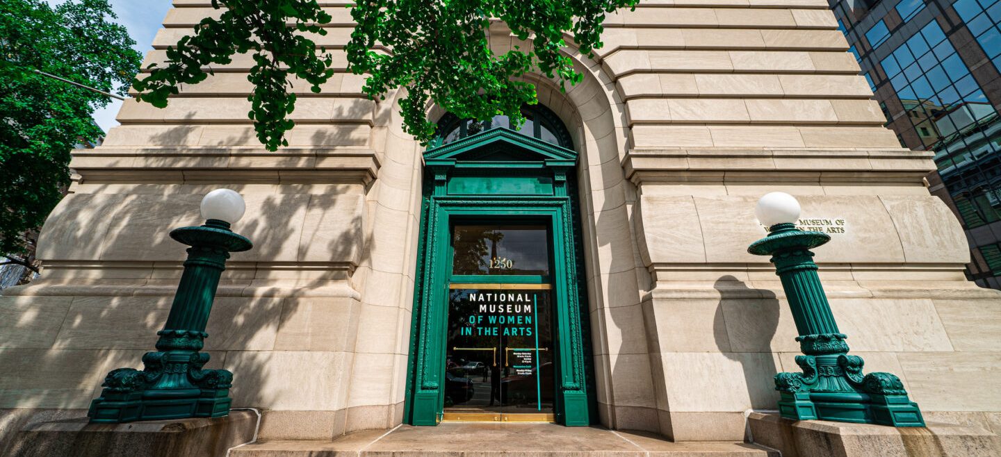 Front-facing photograph of the building exterior with green doors, posts, and tree.