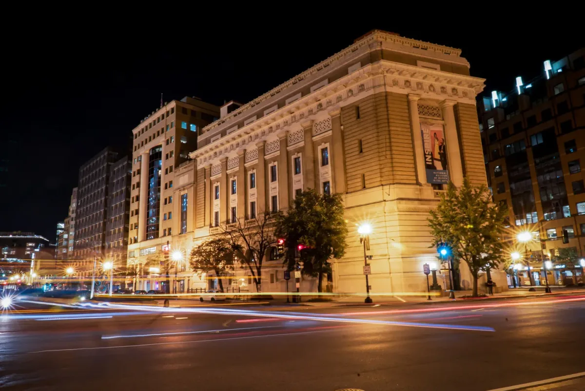 View of the National Museum of Women in the Arts building from outside showing the Neoclassical building from one corner. The building is a tan-colored stone with an arched doorway, long vertical windows, and detailed molding around the roof. It is dark outside, and street lamps shine brightly to illuminate the building.