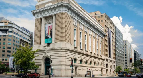 View of the museum from outside showing the Neoclassical building from one corner. The building is a tan-colored stone with an arched doorway, long vertical windows, and detailed molding around the roof.