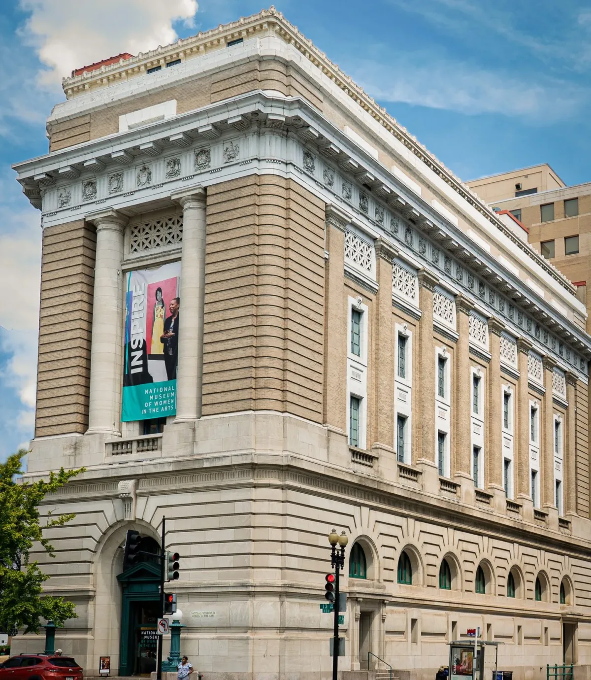 View of the museum from outside showing the Neoclassical building from one corner. The building is a tan-colored stone with an arched doorway, long vertical windows, and detailed molding around the roof.