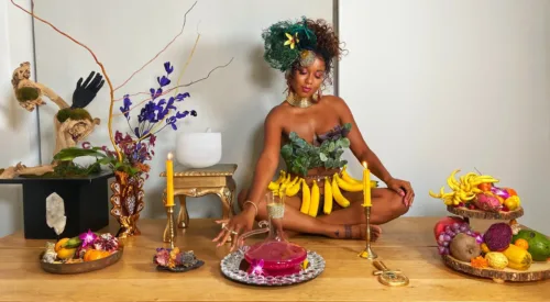 A medium-dark skinned adult woman sits cross-legged on a long wooden table set with platters of colorful fruit, candles, plants, and a pitcher of a pink liquid. She wears green leaves on her chest, gold jewelry, a skirt of bananas, and her hair pulled up with green feathers.