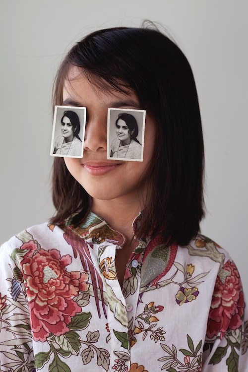 A portrait photograph of a girl with shoulder length straight dark brown hair, wearing a floral-print button-up shirt. She smiles slightly with her lips closed and atop her eyes are two identical black and white portrait photographs of a woman who is in a similar pose as the girl.