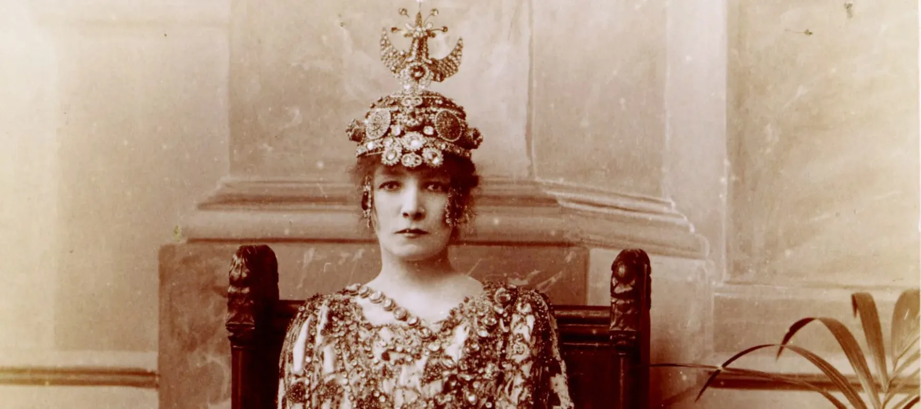 A sepia-toned photograph of a light-skinned woman dressed opulently in a beaded and patterned gown and wearing a beaded, luxurious headpiece.