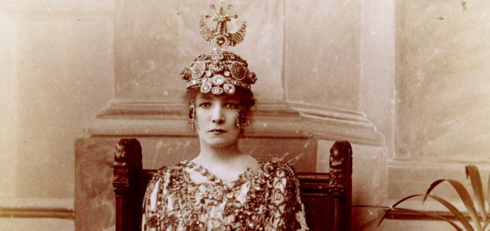 A sepia-toned photograph of a light-skinned woman dressed opulently in a beaded and patterned gown and wearing a beaded, luxurious headpiece.