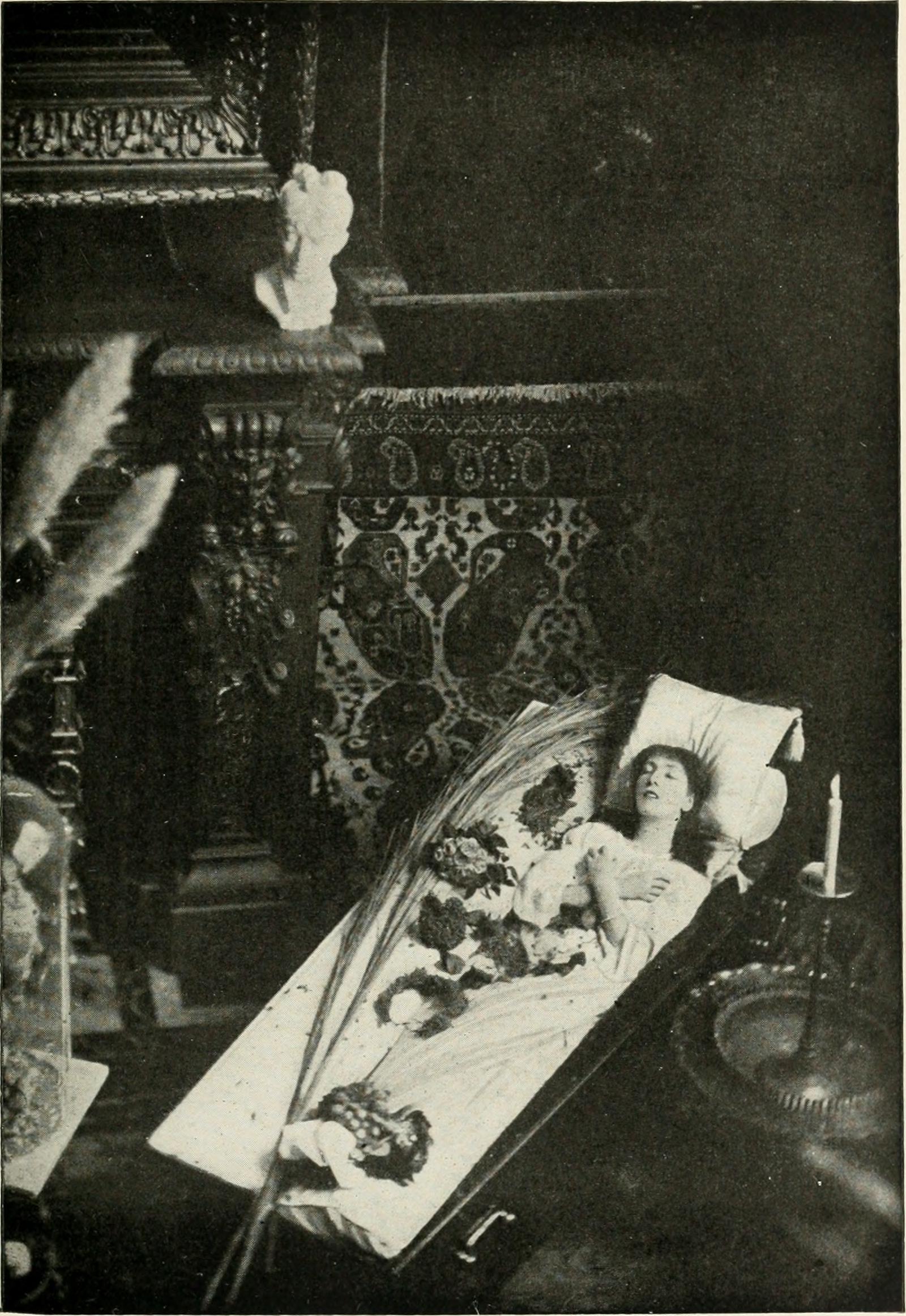 A black-and-white photo of a light-skinned woman laying in a coffin, plush with sheets and a pillow, in a room that had patterned tiled walls, a fireplace atop which a bust sits, and a fancy brass candle holder.