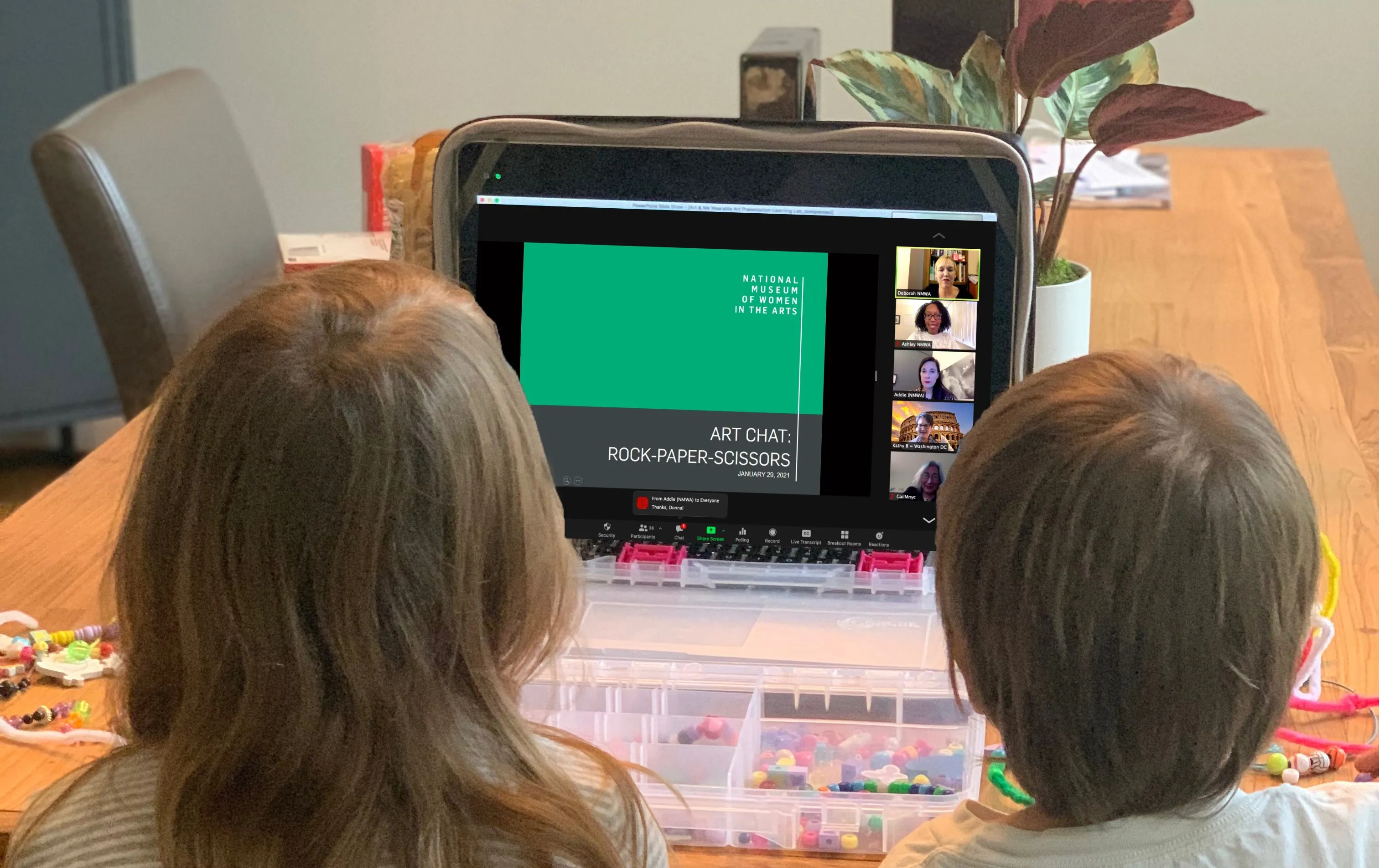 Photograph of the back of two blond children watching an online program on a laptop. The program is titled 'Art Chat: Rock-Paper-Scissors' and is hosted by the National Museum of Women in the Arts.