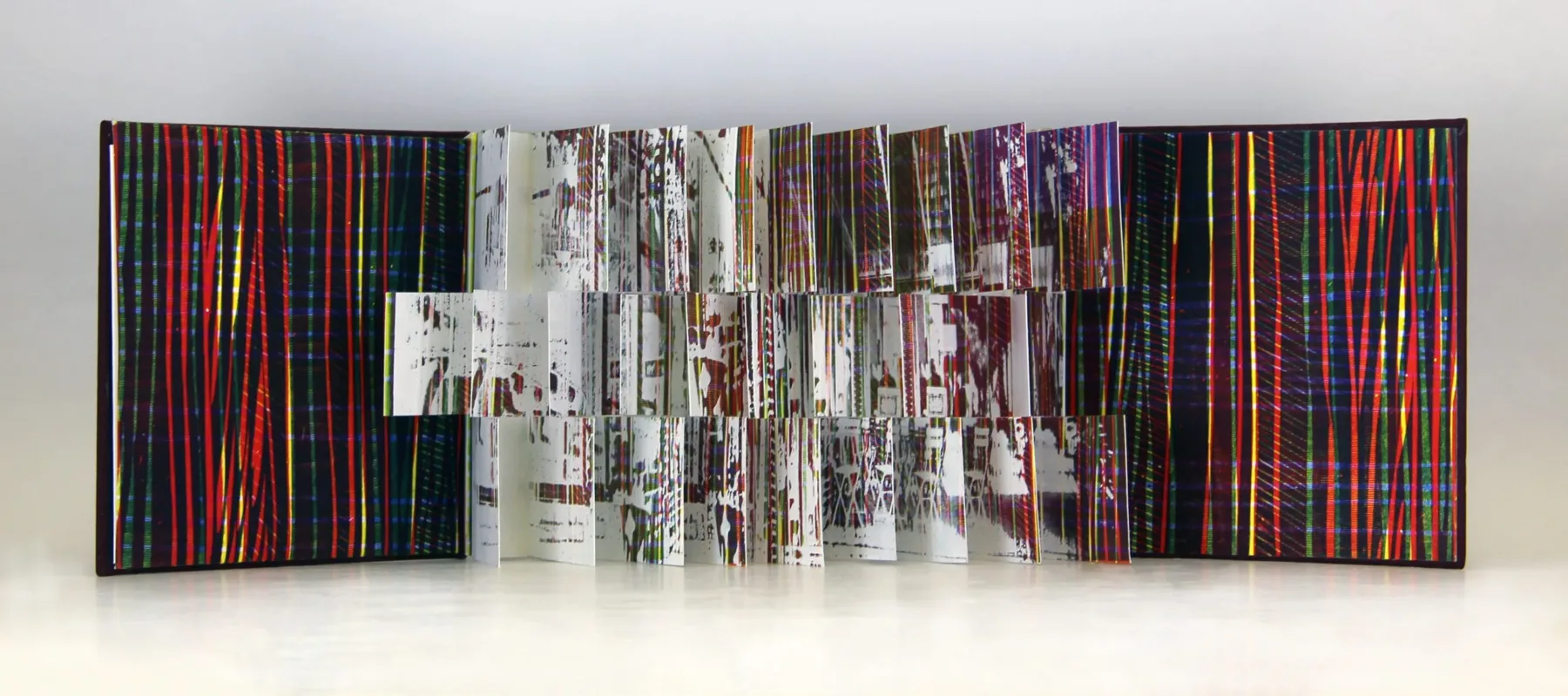 A photograph of an open flag book with red, blue, and yellow diagonal stripes inside the front and back covers. The interior pages are each cut into three sections creating three rows of flags with white splatter paint and multicolored stripes.