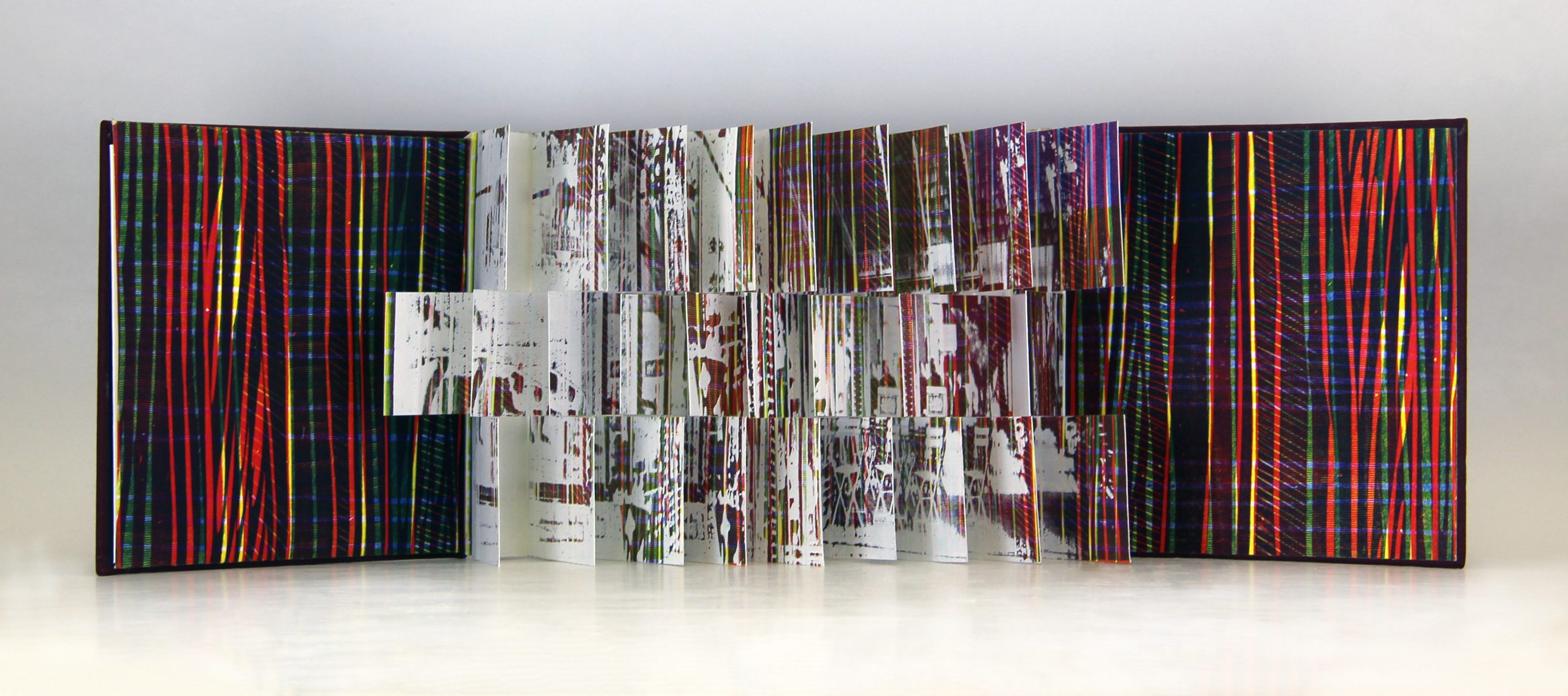 A photograph of an open flag book with red, blue, and yellow diagonal stripes inside the front and back covers. The interior pages are each cut into three sections creating three rows of flags with white splatter paint and multicolored stripes.