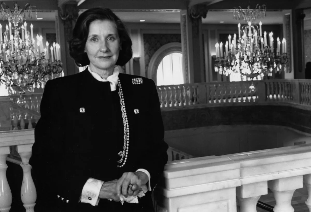 Black-and-white photograph of the museum founder, a white woman in a dark suit, standing on the Mezzanine level of the museum with her hands clasped as she leans against the marble railing in front of the Great Hall chandeliers.