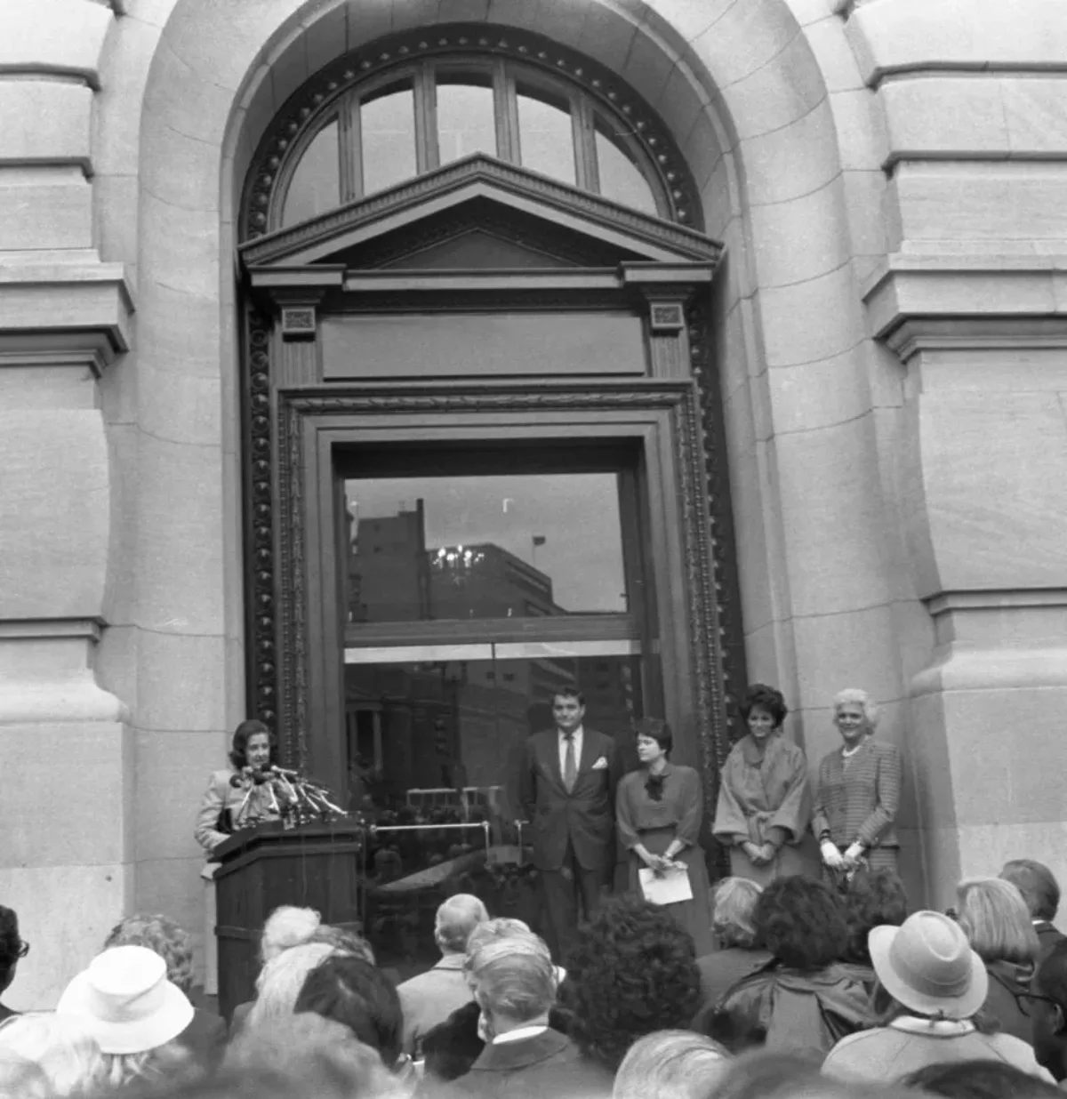 A light-skinned woman speaks at a podium adorned with many microphones outside of a large building. A crowd of people look on and four others, three women and a man, stand in front of the crowd, to the side of the woman with their hands clasped in front of them.