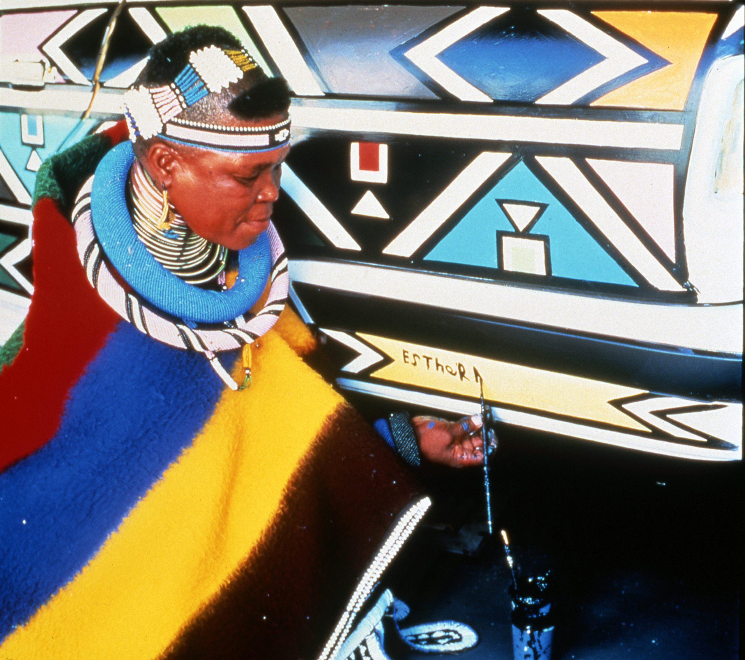 A dark-skinned woman wearing traditional African dress, paints her name on a car featuring colorful, geometric designs.