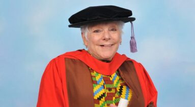 A photographic portrait of smiling art scholar Linda Nochlin in academic regalia. She wears a multicolored blouse and a black cap with a lavender tassel. Over the blouse she wears a bright red and brown gown. She holds a rolled white paper tied with a red ribbon.