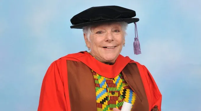 A photographic portrait of smiling art scholar Linda Nochlin in academic regalia. She wears a multicolored blouse and a black cap with a lavender tassel. Over the blouse she wears a bright red and brown gown. She holds a rolled white paper tied with a red ribbon.