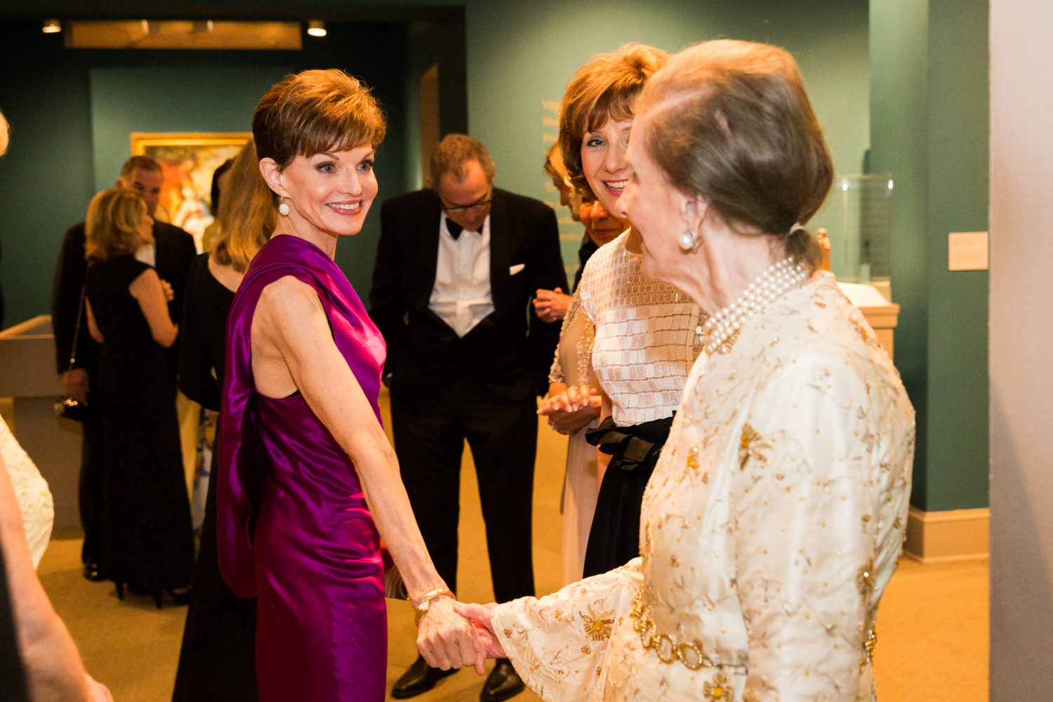 A woman with light skin and brown hair wearing a violet-colored sleeveless dress shakes hands with a woman with light skin wearing a gold dress at a formal party.