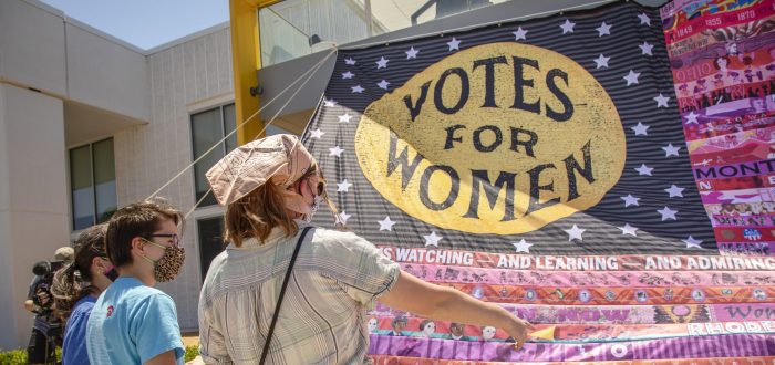Three light-skinned people stand in front of a large textile installation that mimics the U.S. flag. Over the stars square "Votes for Women" is written in a yellow circle. The stripes on the rest of the flag are done in various shades of pink and red and each feature a different theme or imagery, most of which is indistinguishable from the distance the photo is taken.