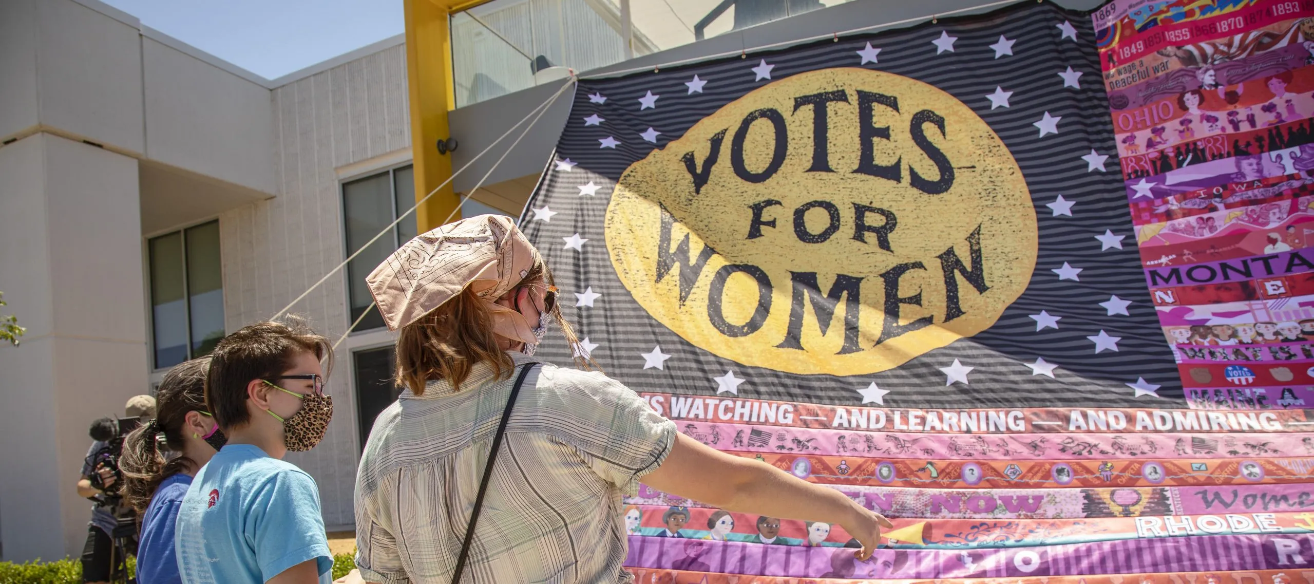 Three light-skinned people stand in front of a large textile installation that mimics the U.S. flag. Over the stars square "Votes for Women" is written in a yellow circle. The stripes on the rest of the flag are done in various shades of pink and red and each feature a different theme or imagery, most of which is indistinguishable from the distance the photo is taken.