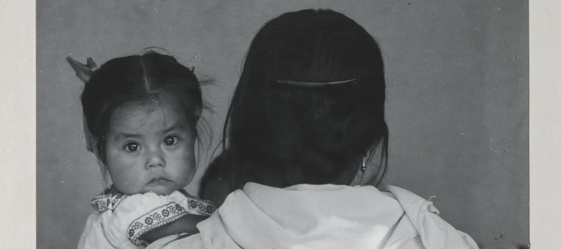 Black-and-white photograph of a woman wearing a dark skirt and light blouse holding a child. The woman faces back and her dark braid reaches past her waist. The dark-haired child stares at the camera and wears a white embroidered dress. Other figures are slightly visible beyond.