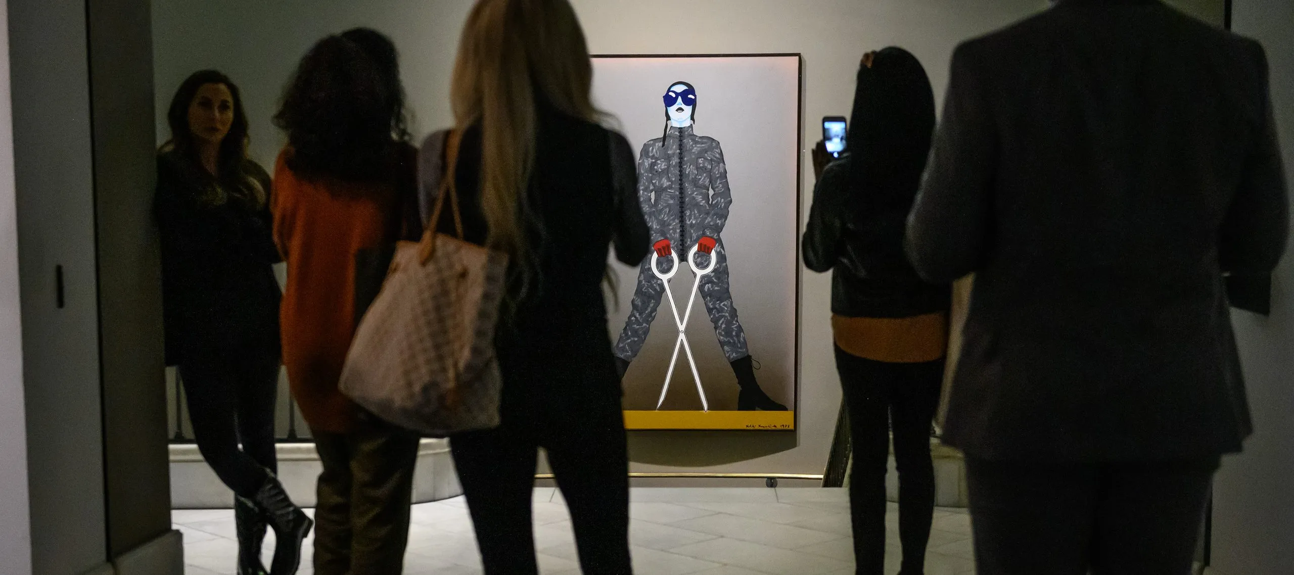 A group gathers in front of a large painting on a woman in camo with a large pair of white scissors.