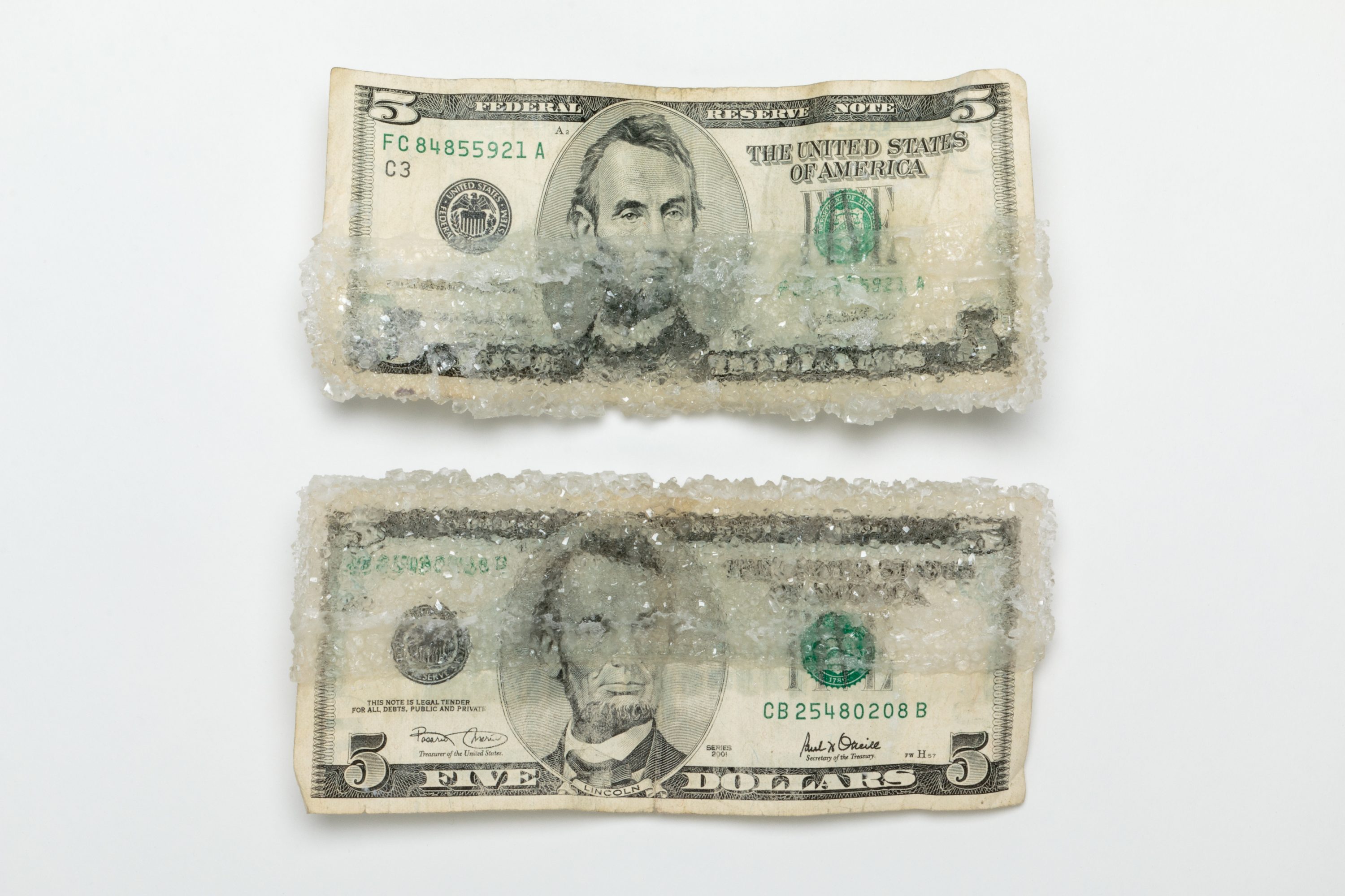 Two five dollar bills lay atop a white background. On the top bill, thick crystals of sugar encrust its lower half; on the bottom bill, thick crystals of sugar encrust its upper half.