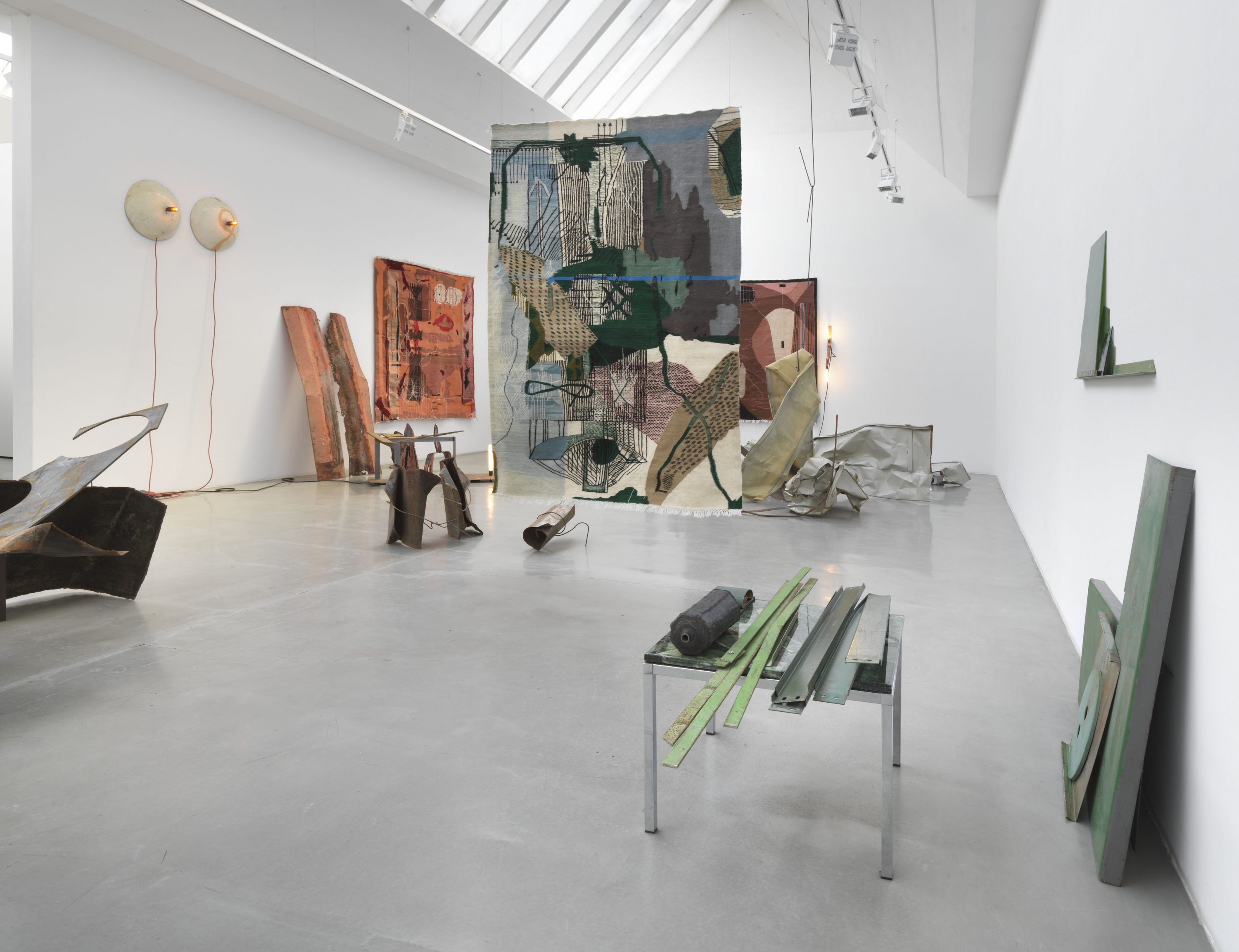 An installation photograph of abstract sculptures in a gallery. White, orange, and gray-toned wooden planks, metal offcuts, and industrial scrap are propped against the white gallery walls and lie on the concrete floor. Multi-colored orange, blue-green, and red tapestries hang on the walls and from the ceiling.