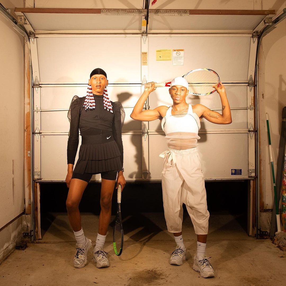 Two dark-skinned people stand in front of a garage door that is open slightly toward the bottom, revealing darkness. One is dressed all black, a tennis skirt, bike shorts, and long sleeve shirt, a du-rag and red and white beaded braids. The other person is dressed in white and khaki pants and a tank top, a white du rag, and holds a tennis racket above their head.