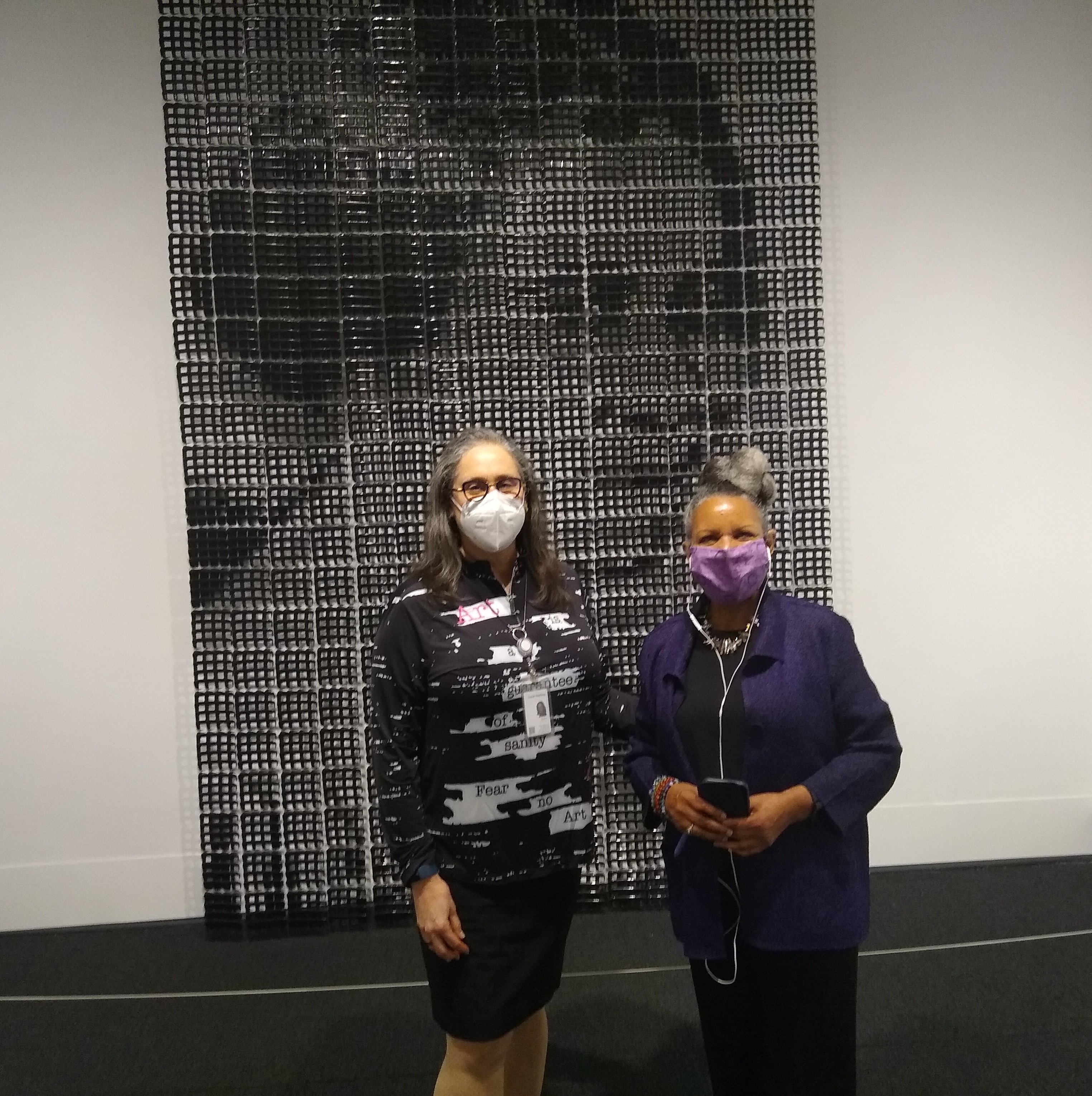 In front of a large portrait of an African American woman made from black plastic pocket combs, a light-skinned woman and dark-skinned woman stand side-by-side smiling behind face masks. The light skinned woman extends her arm behind the other woman's back.
