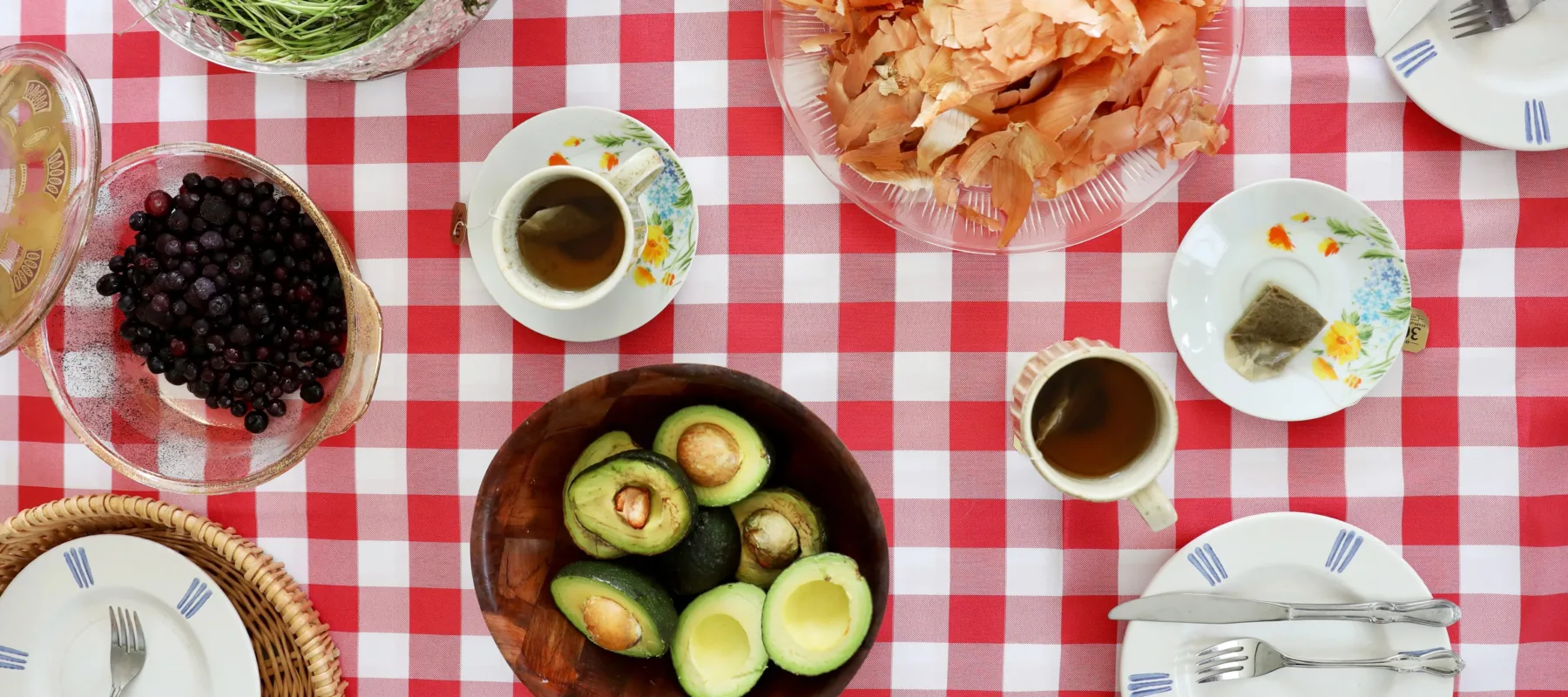 Circular glass, wood, and ceramic plates and bowls arranged on a red and white checkered tablecloth, seen from above. The plates and bowls are filled with blueberries, long carrot stems, halved avocados, an onion and onion peels, and a cups of tea and teabags on saucers.