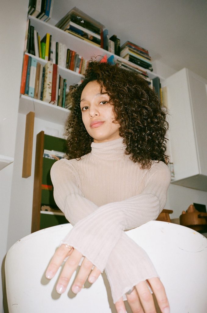 A film photograph taken from a low angle of a light-skinned Black woman with shoulder-length curly hair. She wears a white turtle neck and has her arms extended but crossed at the wrists. She looks at the camera with a slight smile.