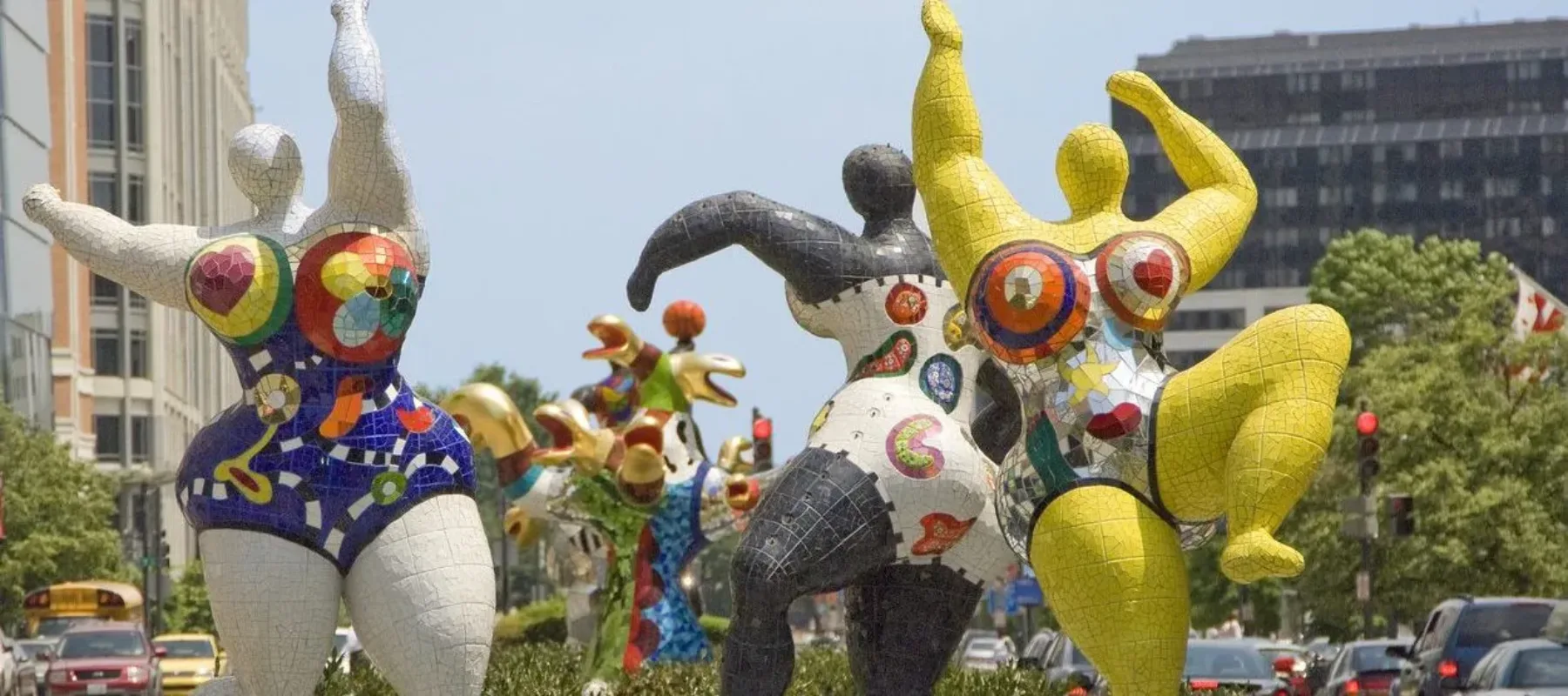 Three abstract outdoor sculptures of voluptuous figures covered in bright patterns and dancing with outstretched arms.