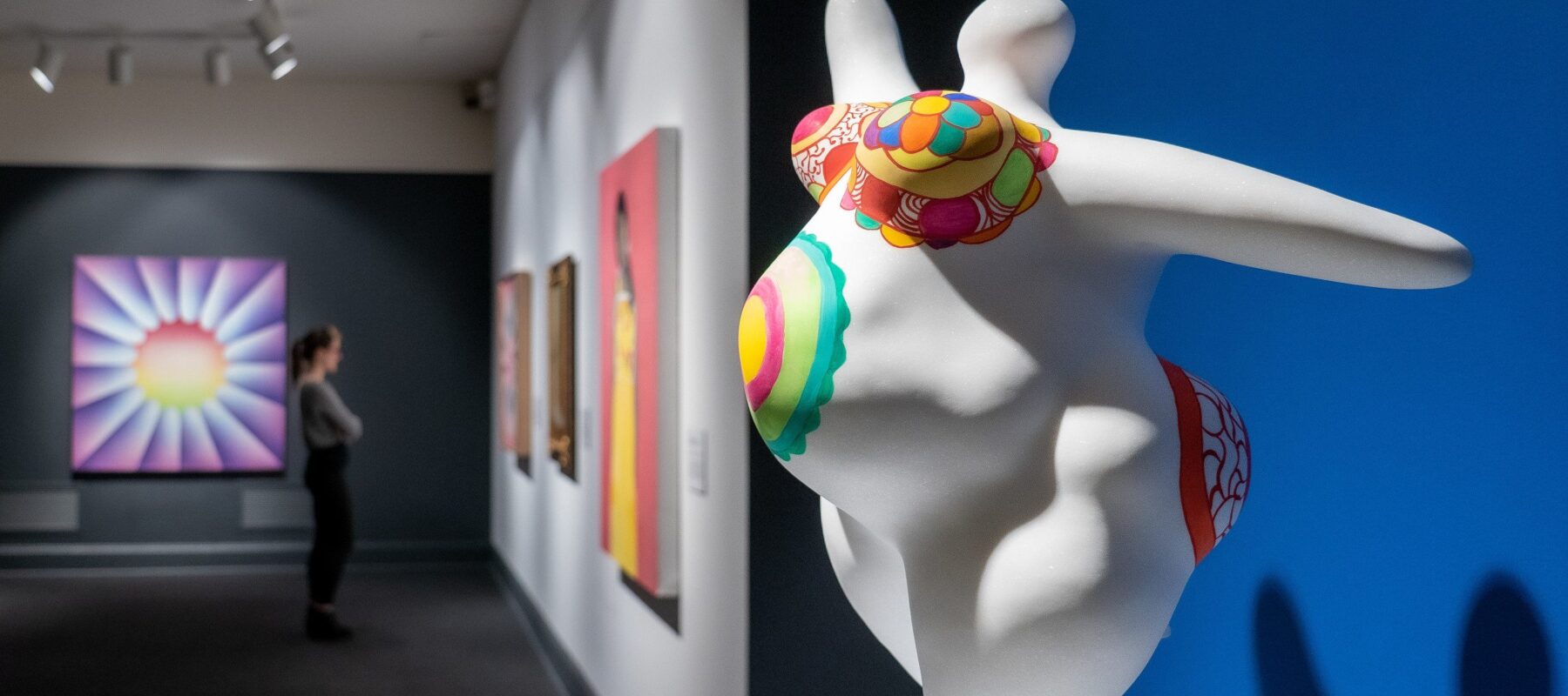 View of the gallery shows an eye-catching marble sculpture in the foreground and a visitor looking at multiple brightly colored artworks in the background. The abstract scupture is of a voluptuous figure with pregnant belly covered in bright patterns and posed with outstretched arms.