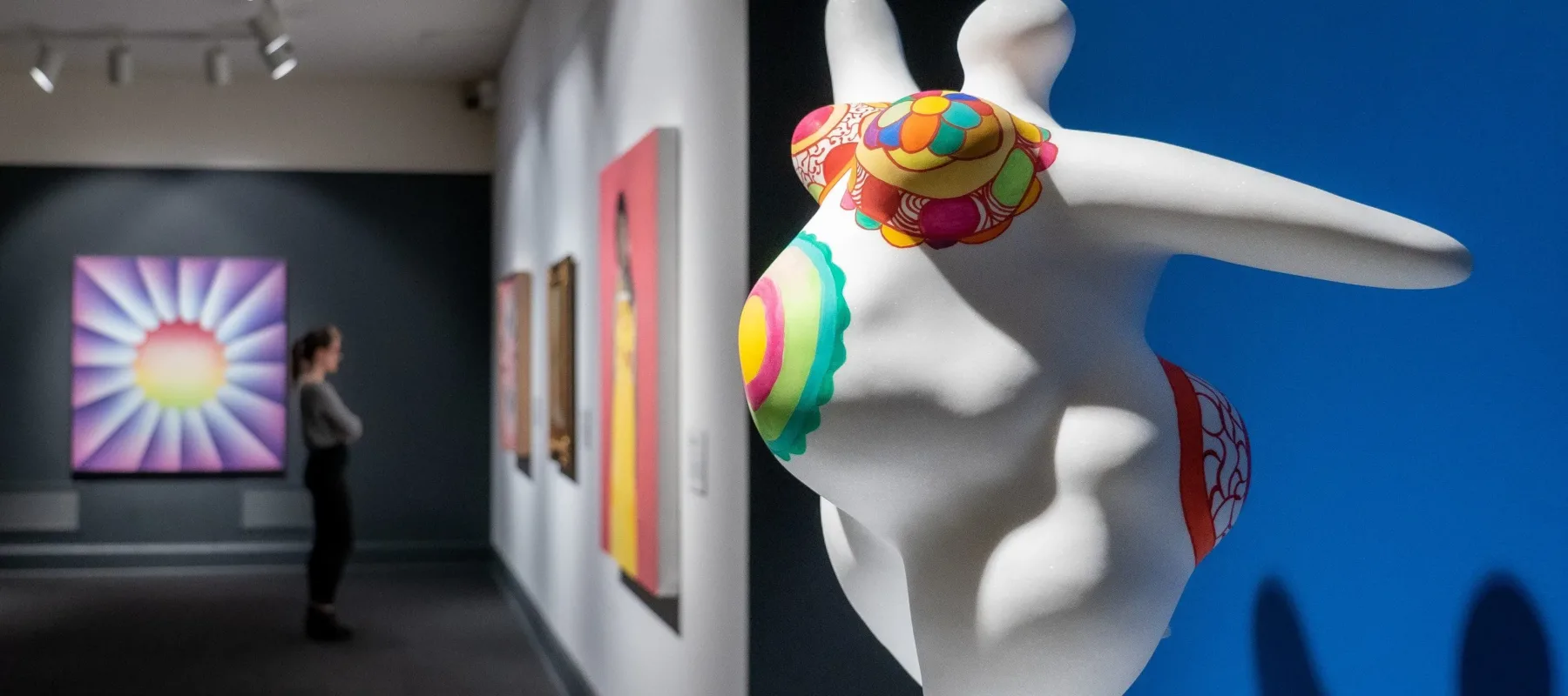 View of the gallery shows an eye-catching marble sculpture in the foreground and a visitor looking at multiple brightly colored artworks in the background. The abstract scupture is of a voluptuous figure with pregnant belly covered in bright patterns and posed with outstretched arms.