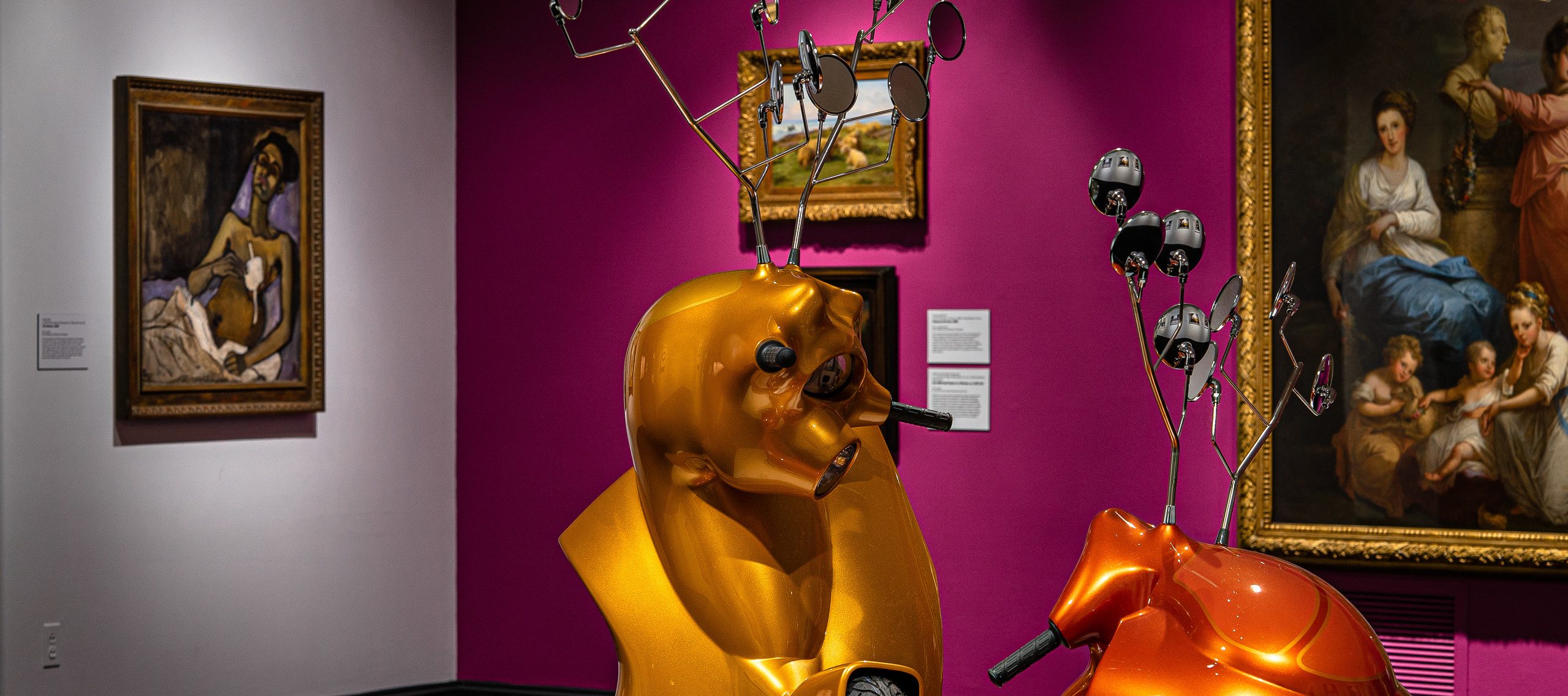 Gallery view with contemporary sculpture made of motorscooters that resemble two antlered animals fighting in foreground.Three paintings hang on a magenta wall in background. To the left on a white wall hangs another painting.