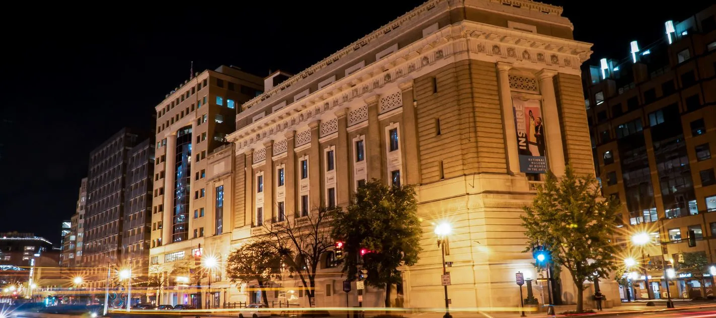 View of the National Museum of Women in the Arts building from outside showing the Neoclassical building from one corner. The building is a tan-colored stone with an arched doorway, long vertical windows, and detailed molding around the roof. It is dark outside, and street lamps shine brightly to illuminate the building.