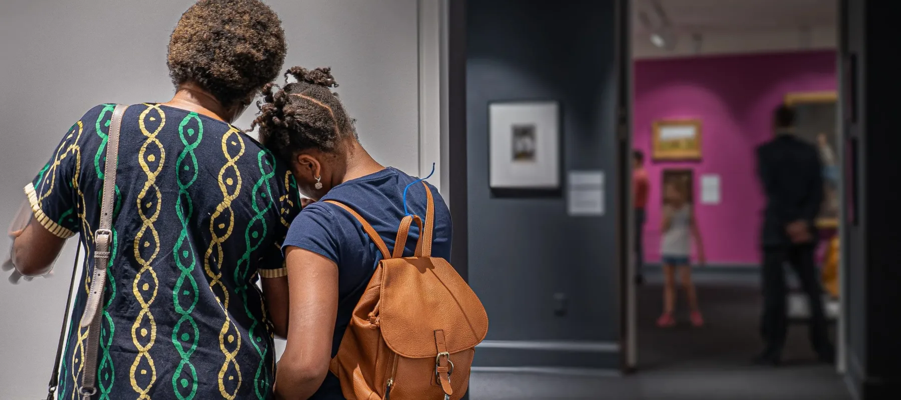 A middle aged, medium dark-skinned woman and a school age medium dark skinned girl face away from the camera and lean into each other affectionately while viewing artwork.