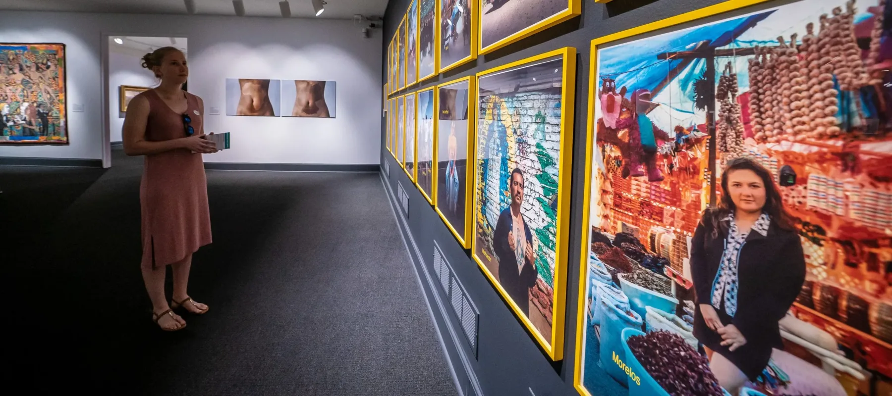 Gallery view of a person with light skin looking at a wall of photographs in bright yellow frames. Two artworks hand on the far wall.