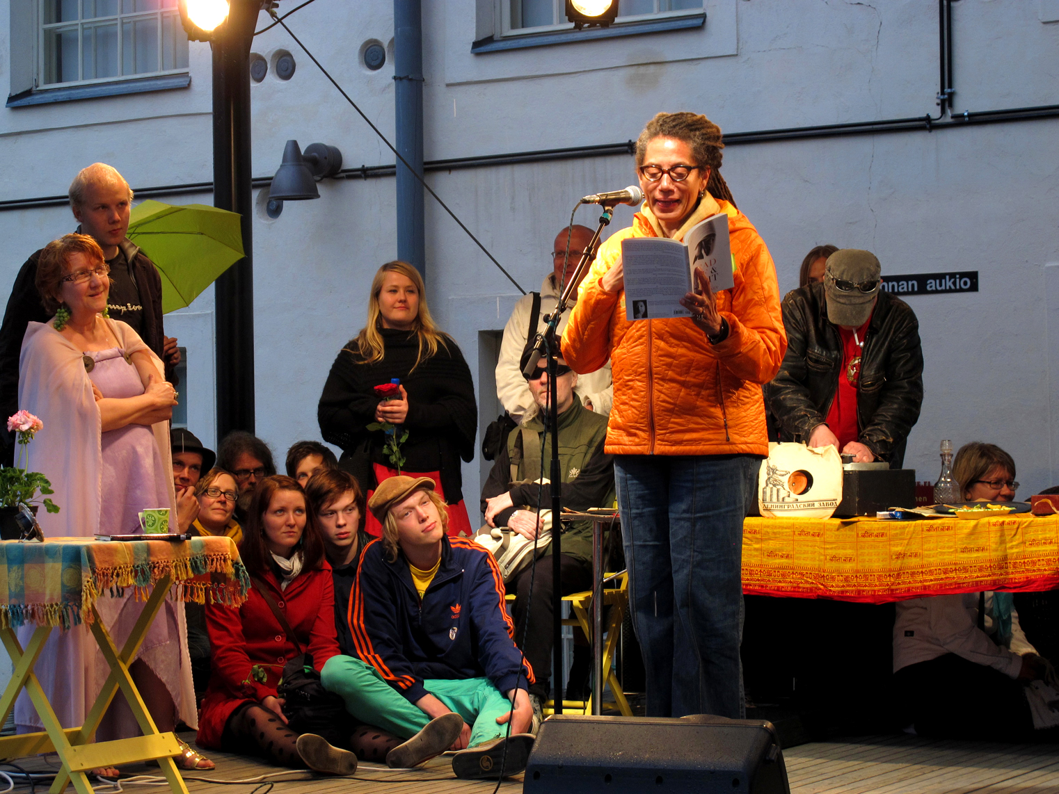 A light-skinned African American woman wears a puffy orange vest and stands in front of a microphone reading from a book. Her long, salt-and-pepper dreadlocks are pulled back into a ponytail. Behind her, young white people sit on the floor and stand. Listening at attention. 
