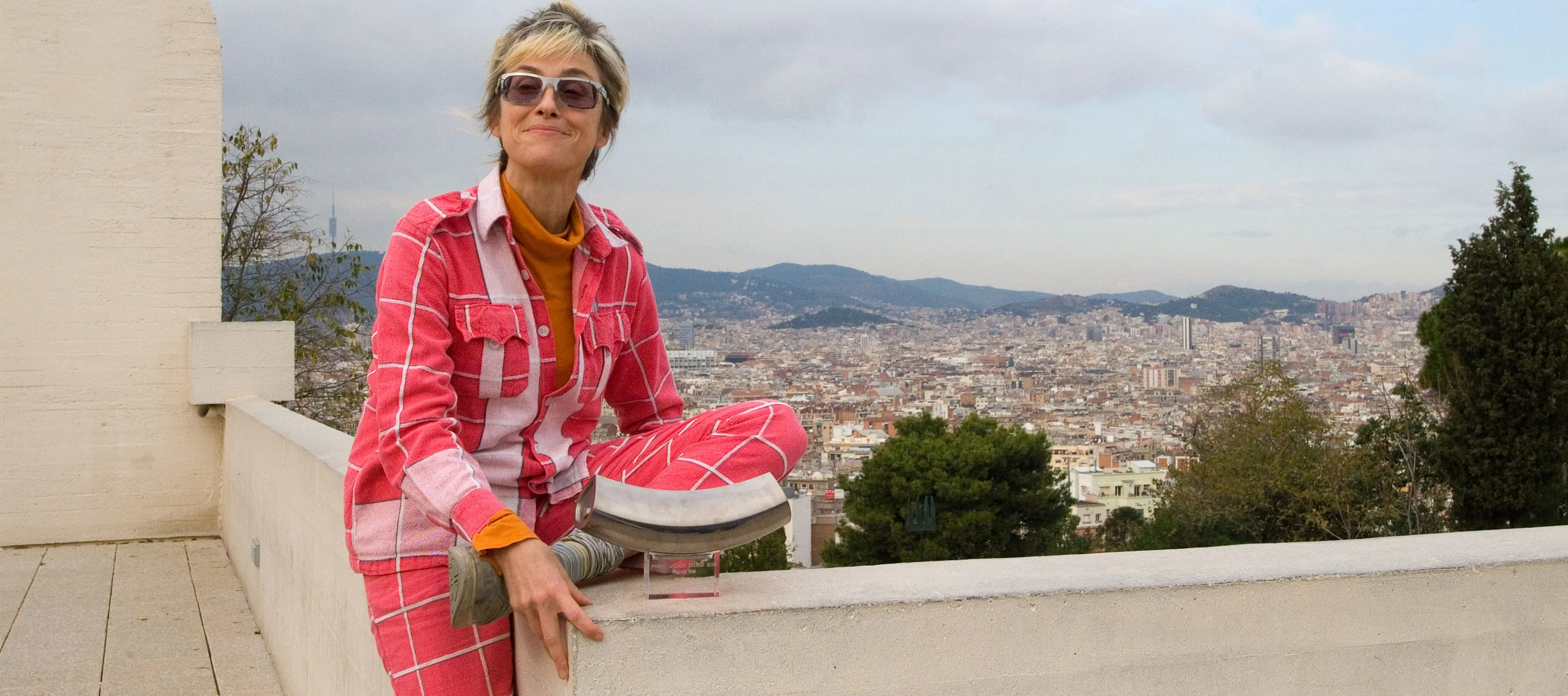 A light-skinned woman with a blonde and brown pixie cut sits atop a cement wall that overlooks Barcelona. She wears a dark and light pink cardigan and pants with checkered white lines over an orange turtleneck. She sits casually on the wall with one left folded under her and the other long, touching the ground. She smiles mischievously at the camera.