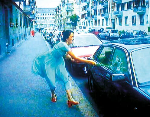 A light-skinned woman in a flowing blue dress and red heels smashes a car window with a slim metal club. Her mouth is open in effort as she swings.