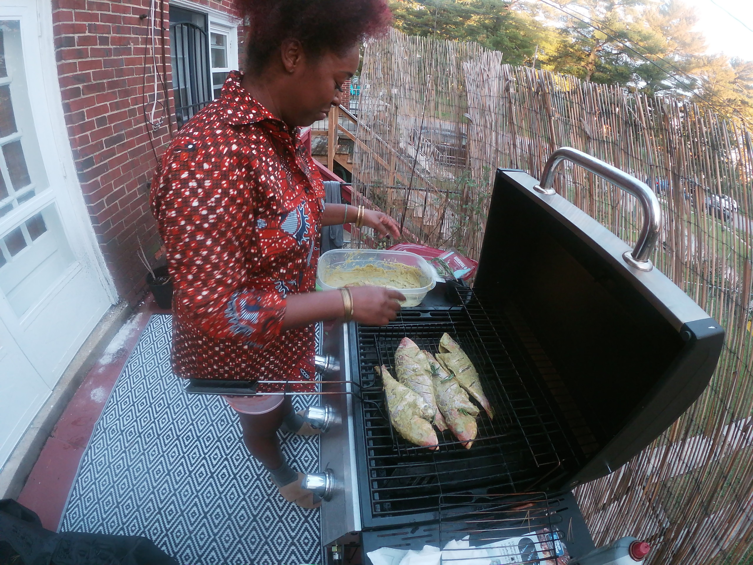 A dark-skinned woman stands on an outdoor balcony, grilling four whole fish on a gas grill. She holds a Tupperware of marinade. The photo is taken with a GoPro, creating a fish eye angle of the scene.