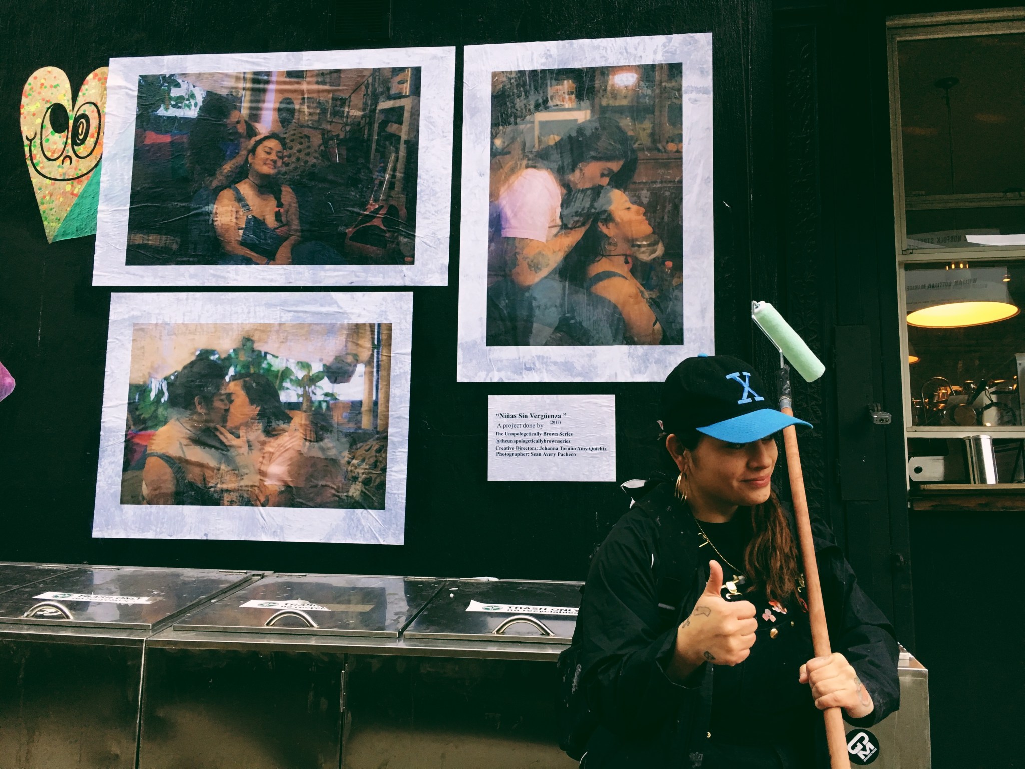 A medium-skinned woman wearing a black baseball cap with a blue "X" on it stands in front of several photographs wheatpasted to a dark wall. She give the camera a thumbs up.