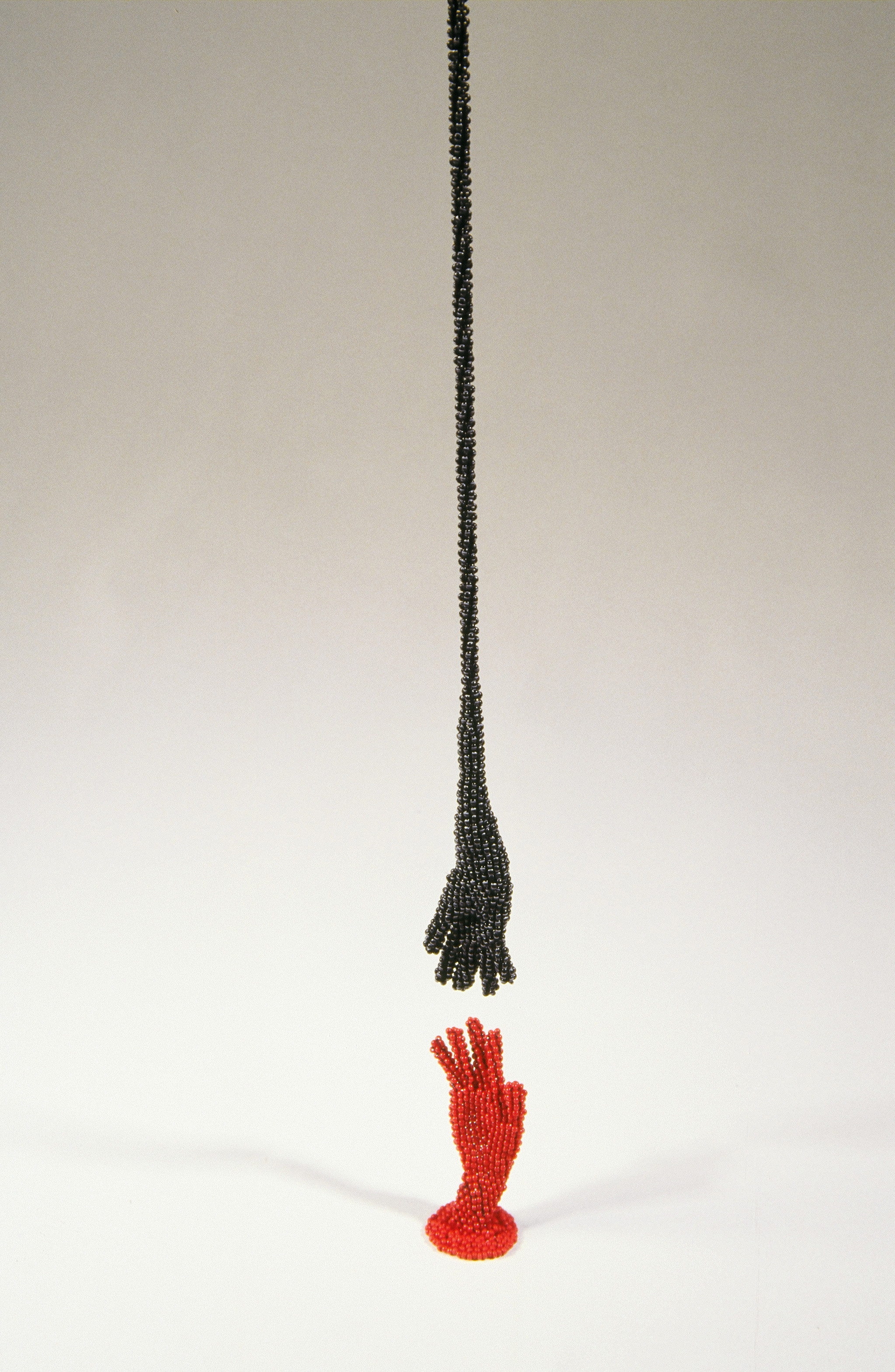 Two small hands are made from tiny black and red beads. The red hand stands upright on a flat white surface, A black hand with a long 
