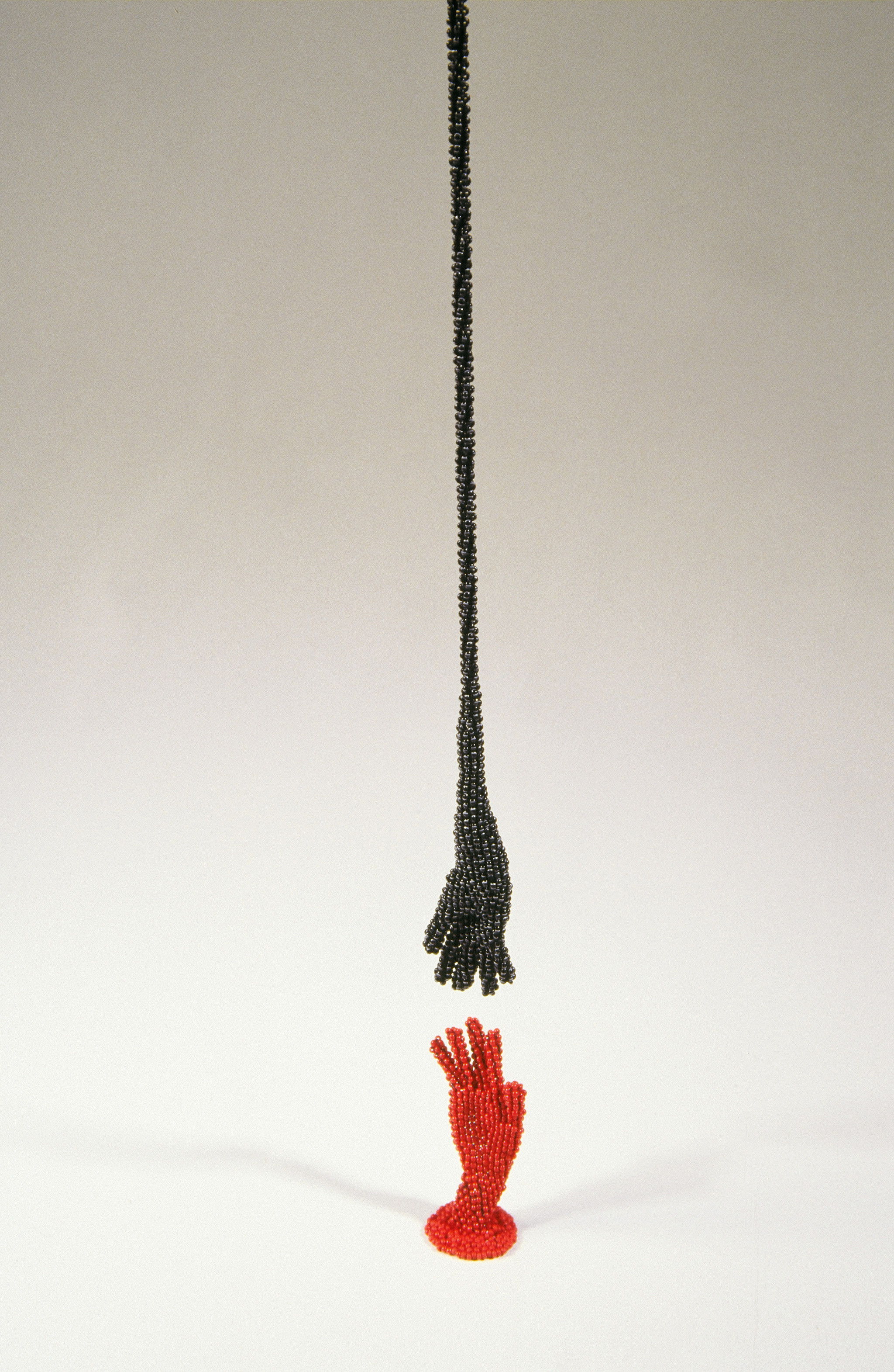 Two small hands are made from tiny black and red beads. The red hand stands upright on a flat white surface, A black hand with a long "arm" hangs from above, hovering just above the red hand, as if they are both about to touch.