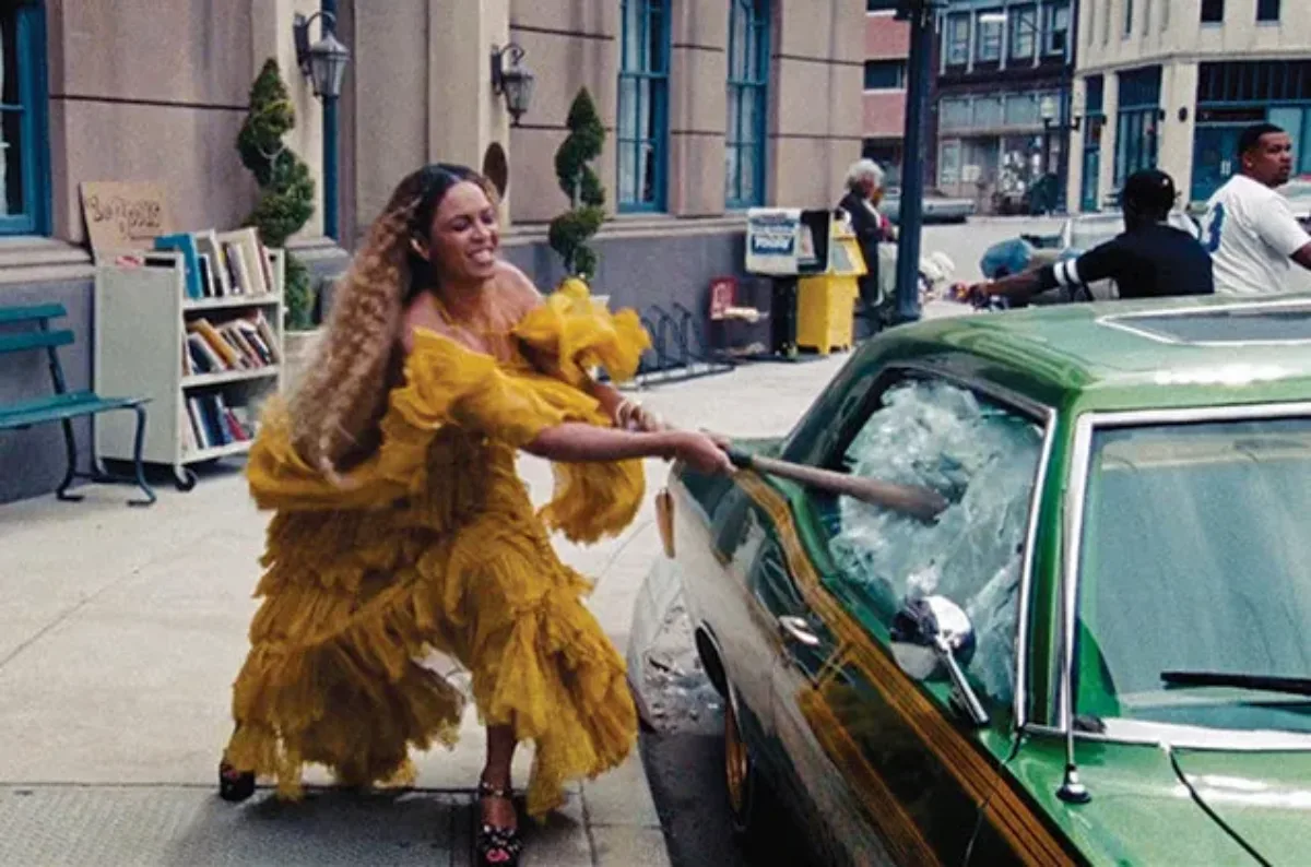 A dark-skinned woman with long, crimped ombre hair smashes a car window with a baseball bat. She wears a flowing, ruffled yellow dress and platform black heels. Her face is focused in a grimace.