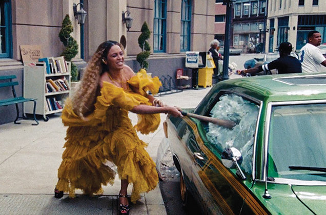 A dark-skinned woman with long, crimped ombre hair smashes a car window with a baseball bat. She wears a flowing, ruffled yellow dress and platform black heels. Her face is focused in a grimace.